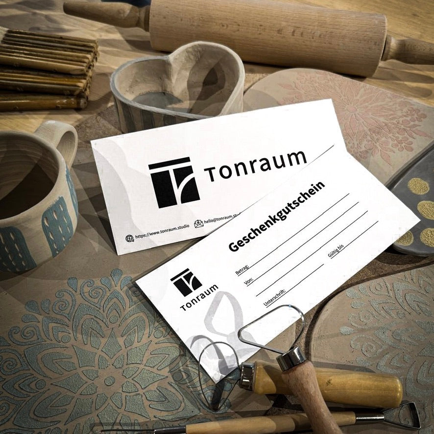 Zwei Tonraum-Geschenkkarten liegen auf einem Tisch mit Töpfermotiven. Im Hintergrund sind Tonwerkzeuge, eine gemusterte Unterlage und Keramikstücke zu sehen. Die Geschenkkarten zeigen das Tonraum-Logo und bieten Platz für weitere Details.