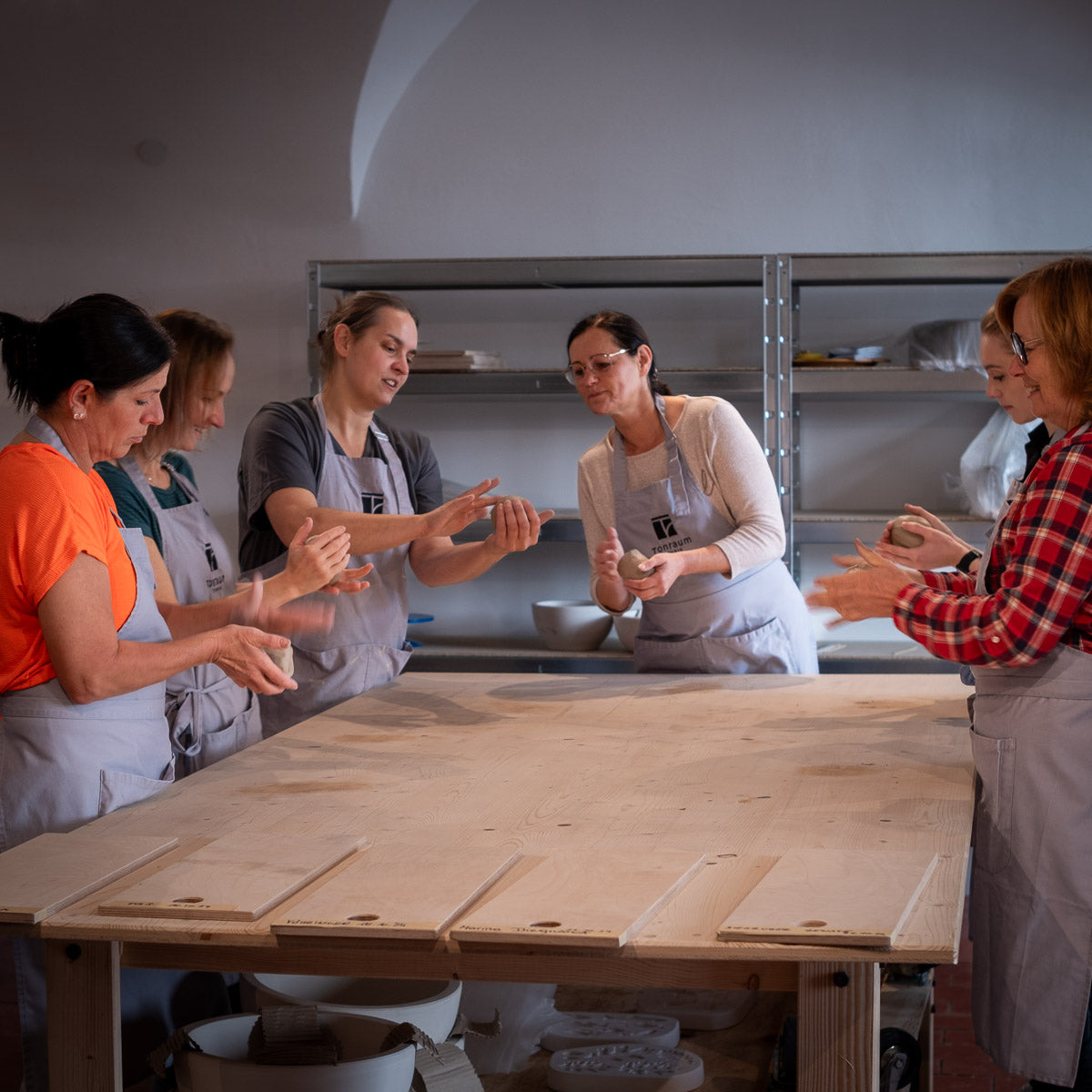 Sechs Frauen in Schürzen formen während einer Töpfersession im Freiraum Ton an einem Holztisch, hinter ihnen Regale voller Töpferwaren und Werkzeuge – und genießen die kreativen Pausen, die das Monatsabo Studionutzung ermöglicht.