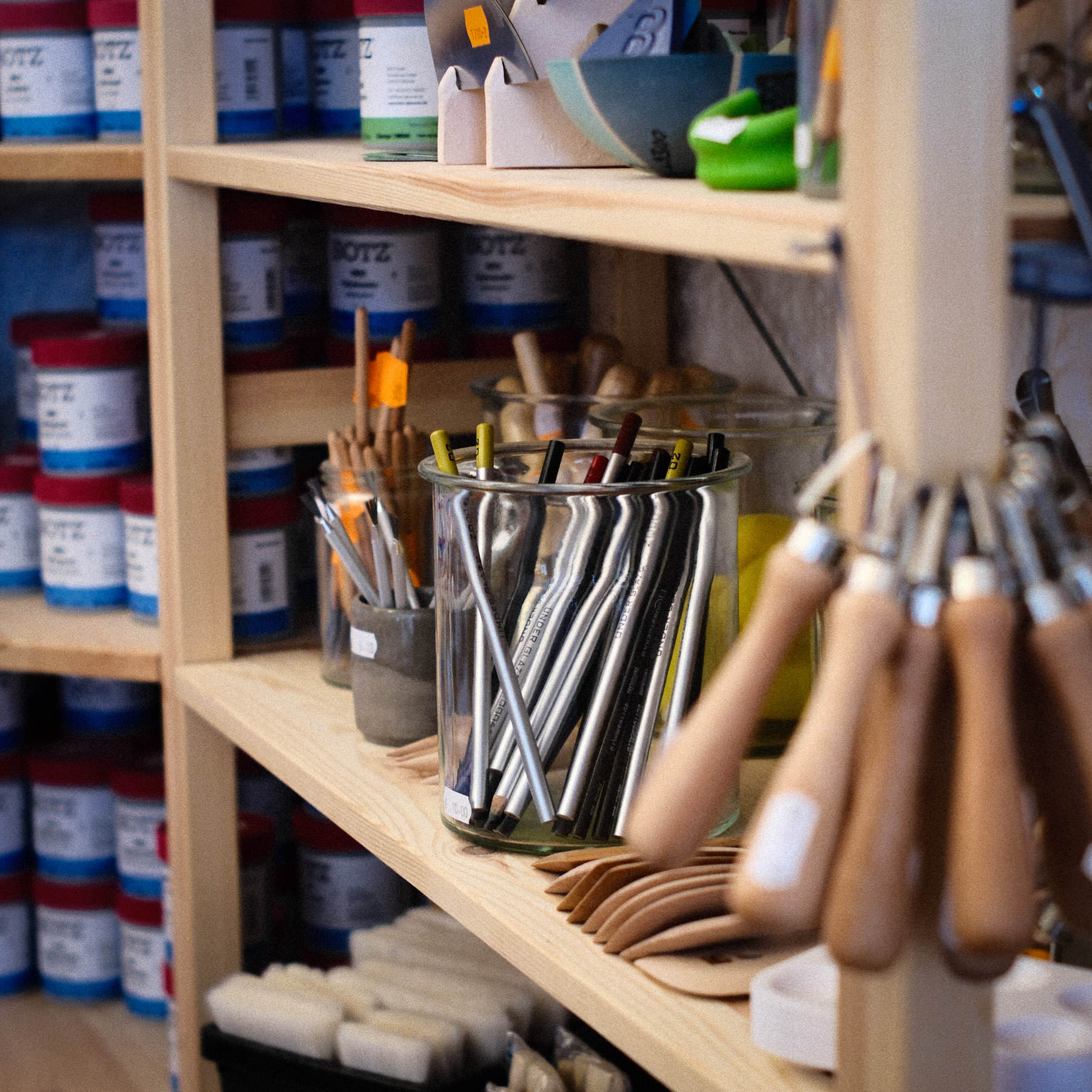 In einem Atelier sind die Regale mit Künstlerbedarf gefüllt, darunter Farbtöpfe, Behälter mit Pinseln und Werkzeugen sowie verschiedene Instrumente mit Holzgriffen.