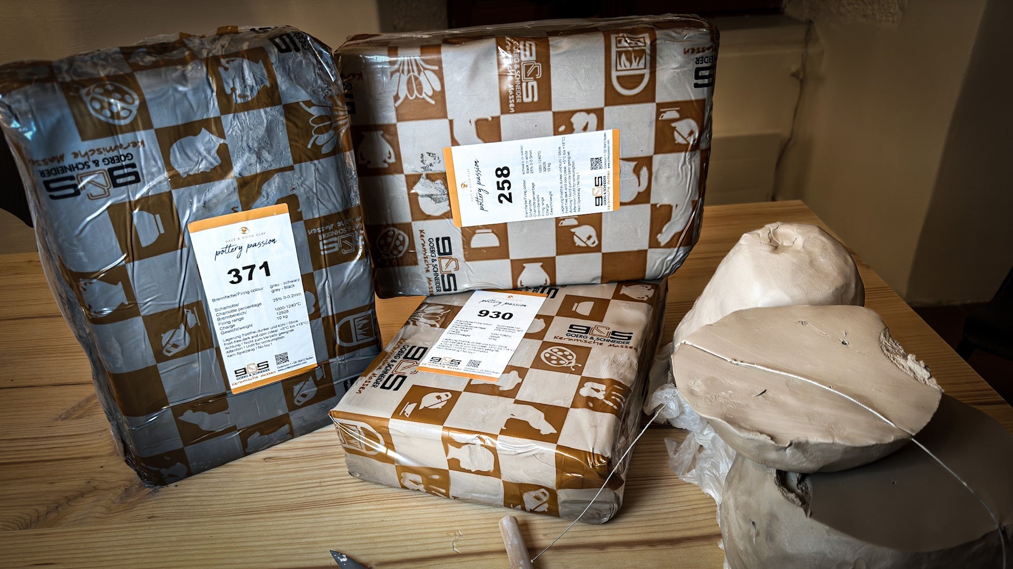 Drei versiegelte Verpackungen mit Etiketten liegen auf einem Holztisch, daneben ein offener Tonblock, einige Drähte und ein Bastelmesser.