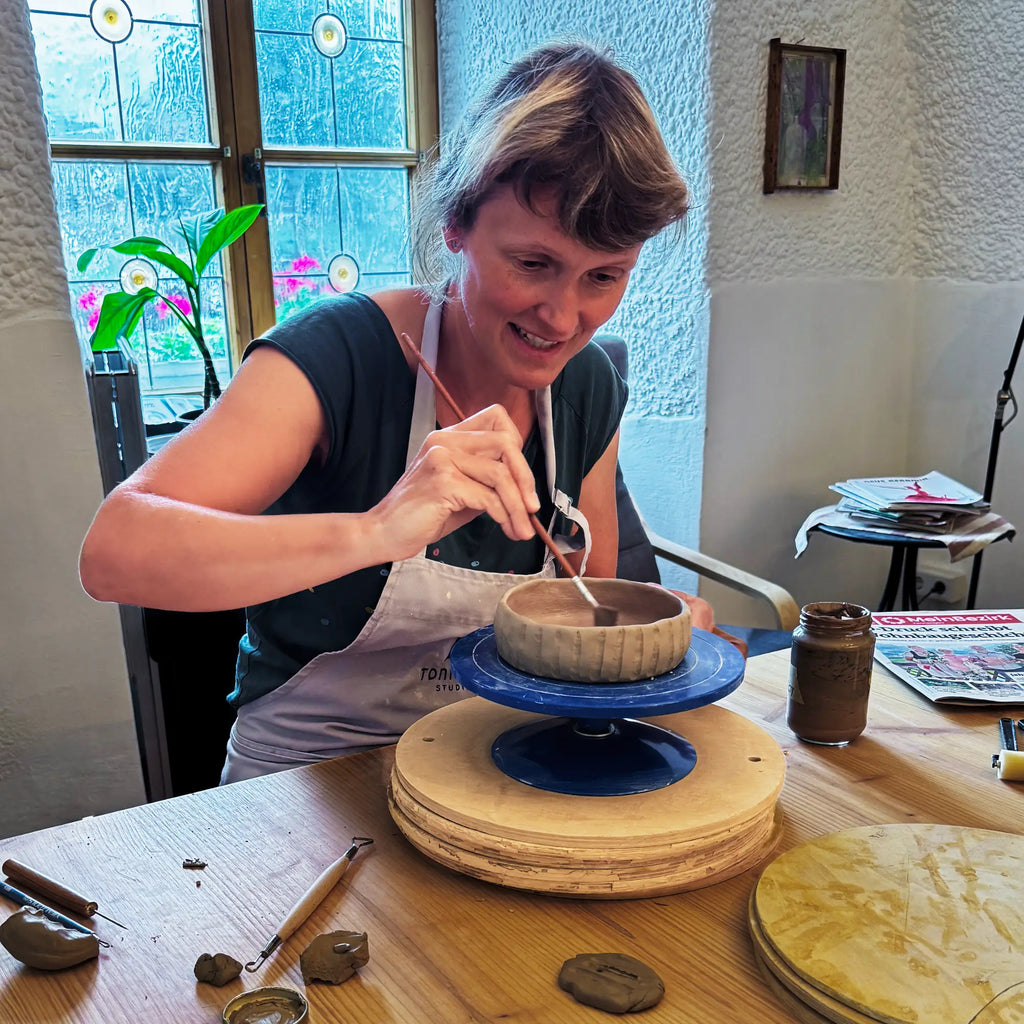 Eine Frau lächelt, als sie während des Afterwork Töpferns eine Keramikschale auf einer Töpferscheibe verziert. Auf dem Holztisch liegen Bildhauerwerkzeuge, Tonstücke und ein Gefäß, während Buntglasfenster ihre kreative Auszeit unterstreichen.
