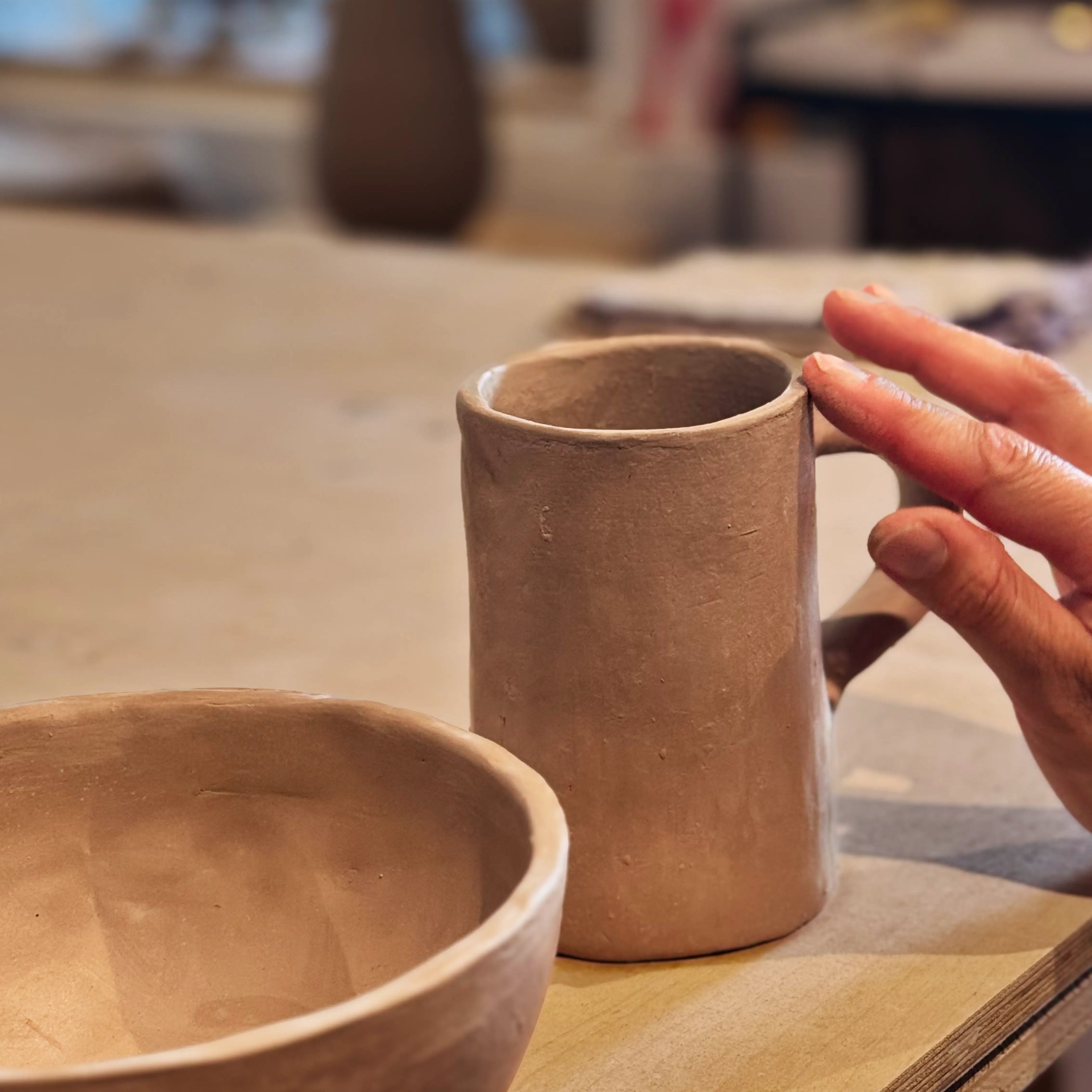 Eine Hand berührt den Griff eines handgefertigten, unglasierten Keramikbechers neben einer unglasierten Tonschale - die perfekte Inspiration für deine kreative Auszeit beim Afterwork Töpfern.