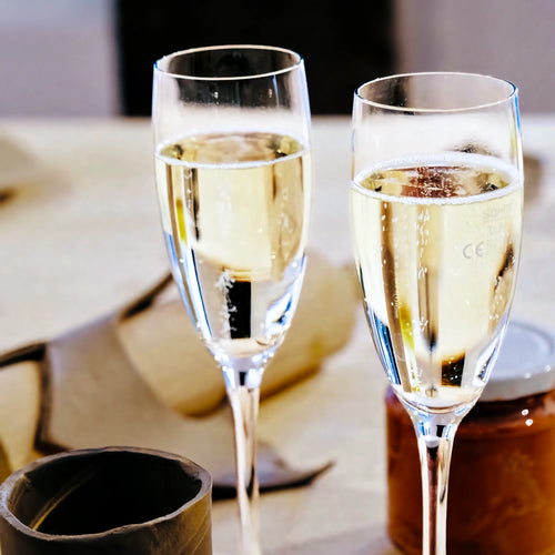 Zwei gefüllte Sektflöten stehen auf einem gedeckten Tisch, daneben liegen eine braune Serviette und ein Glas, was darauf schließen lässt, dass Freunde einen gemeinsamen Afterwork-Töpferabend genießen.