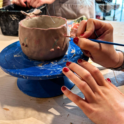 Beim Afterwork Töpfern verziert eine Person mit rotem Nagellack einen handgefertigten Tonbecher auf einer blauen Töpferscheibe, während im Hintergrund eine weitere Person mit Töpferwerkzeugen erscheint.