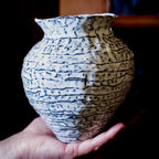 Eine Hand hält eine strukturierte Keramikvase, die mit "Feine Keramik von Hand formen - dein Einstieg in die Aufbautechnik" hergestellt wurde und blaue Wellenlinien und eine geriffelte Oberfläche vor einem dunklen Hintergrund aufweist.