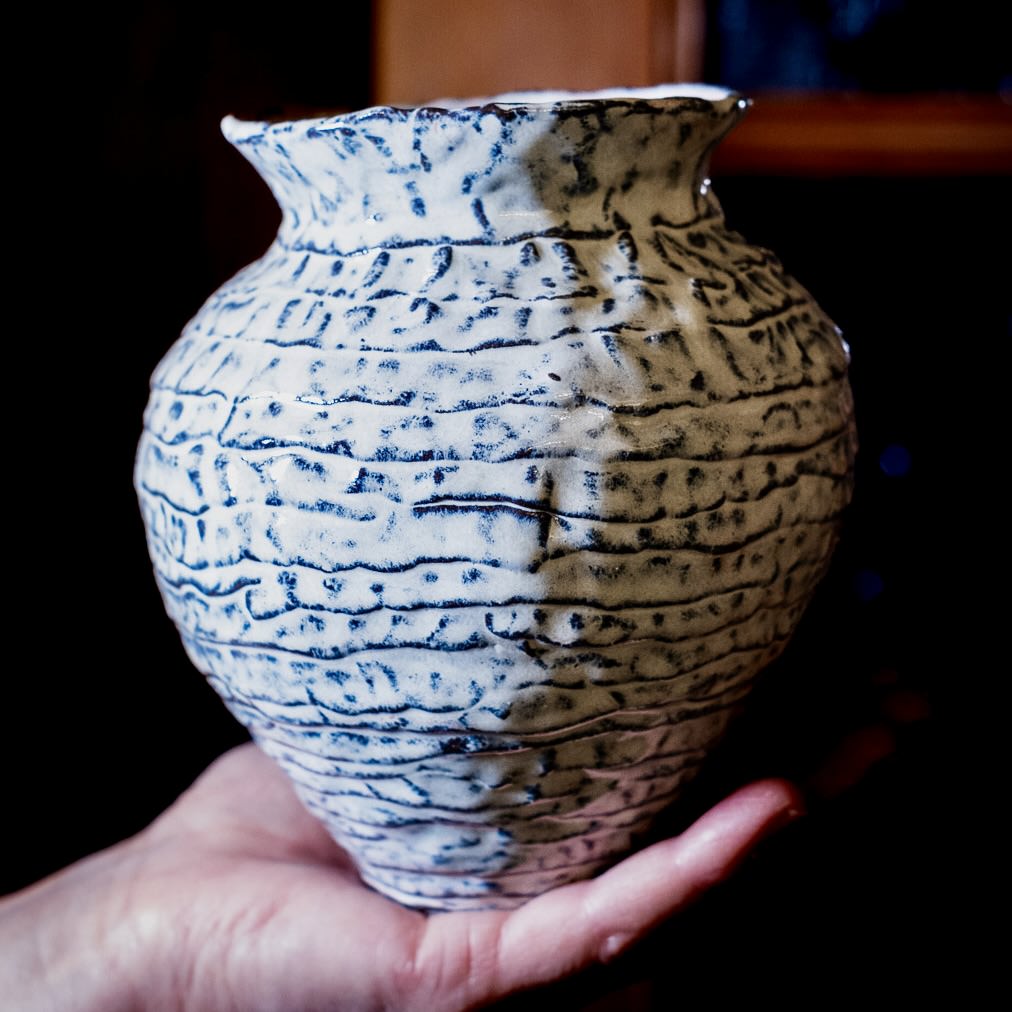 Eine Hand hält eine strukturierte Keramikvase, die mit "Feine Keramik von Hand formen - dein Einstieg in die Aufbautechnik" hergestellt wurde und blaue Wellenlinien und eine geriffelte Oberfläche vor einem dunklen Hintergrund aufweist.