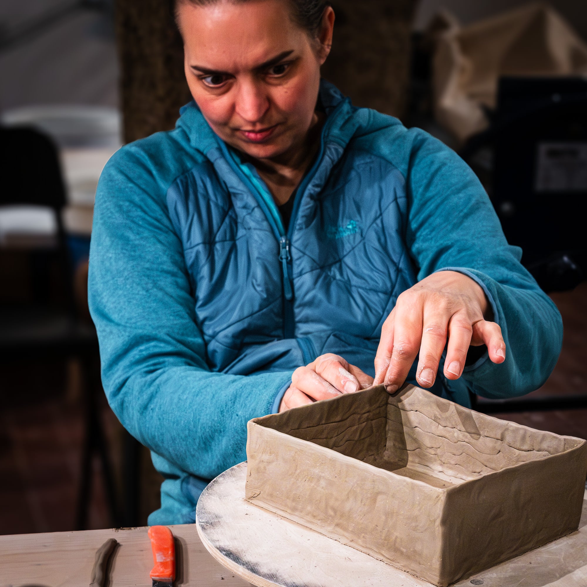 Bei "Feine Keramik von Hand formen - dein Einstieg in die Aufbautechnik" formt eine Frau in einer blauen Jacke gekonnt eine rechteckige Tonschachtel von Hand. Töpferwerkzeuge und Tonreste liegen auf dem Tisch, während sie sich auf ihre Technik konzentriert.