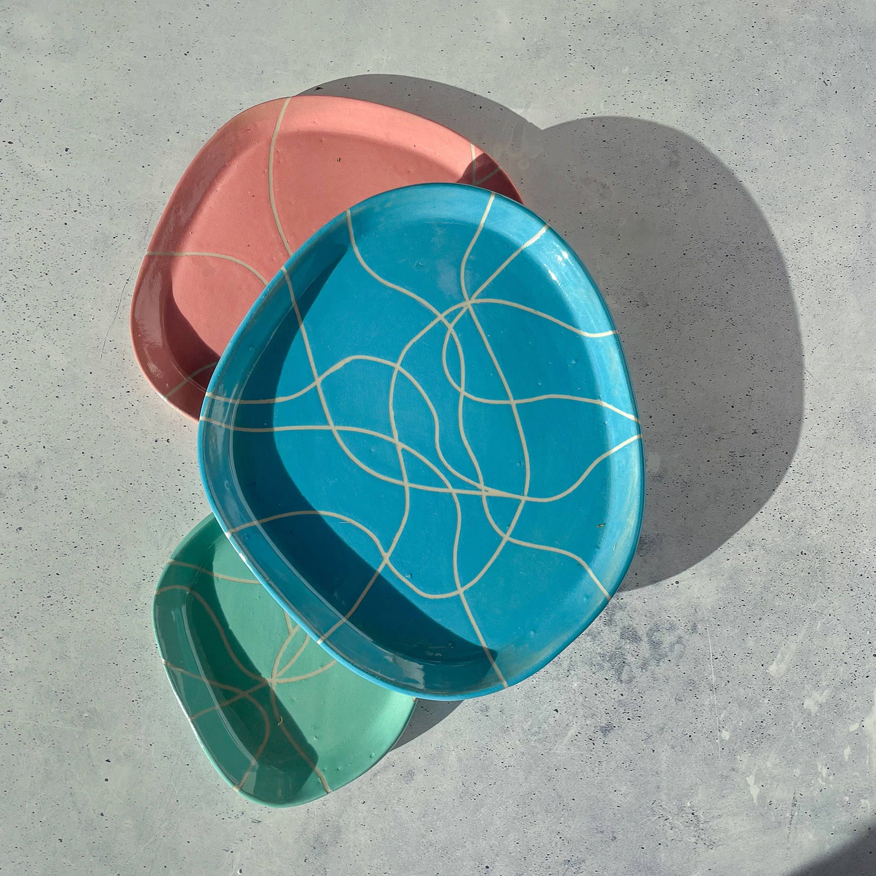 Drei unregelmäßige Keramikteller der Botz Edition - rosa, blau und grün - überlappen sich auf einer hellgrauen, strukturierten Oberfläche. Jeder Teller zeigt abstrakte cremefarbene Linien und eine matte Oberfläche. Das Sonnenlicht wirft weiche Schatten auf die Teller.