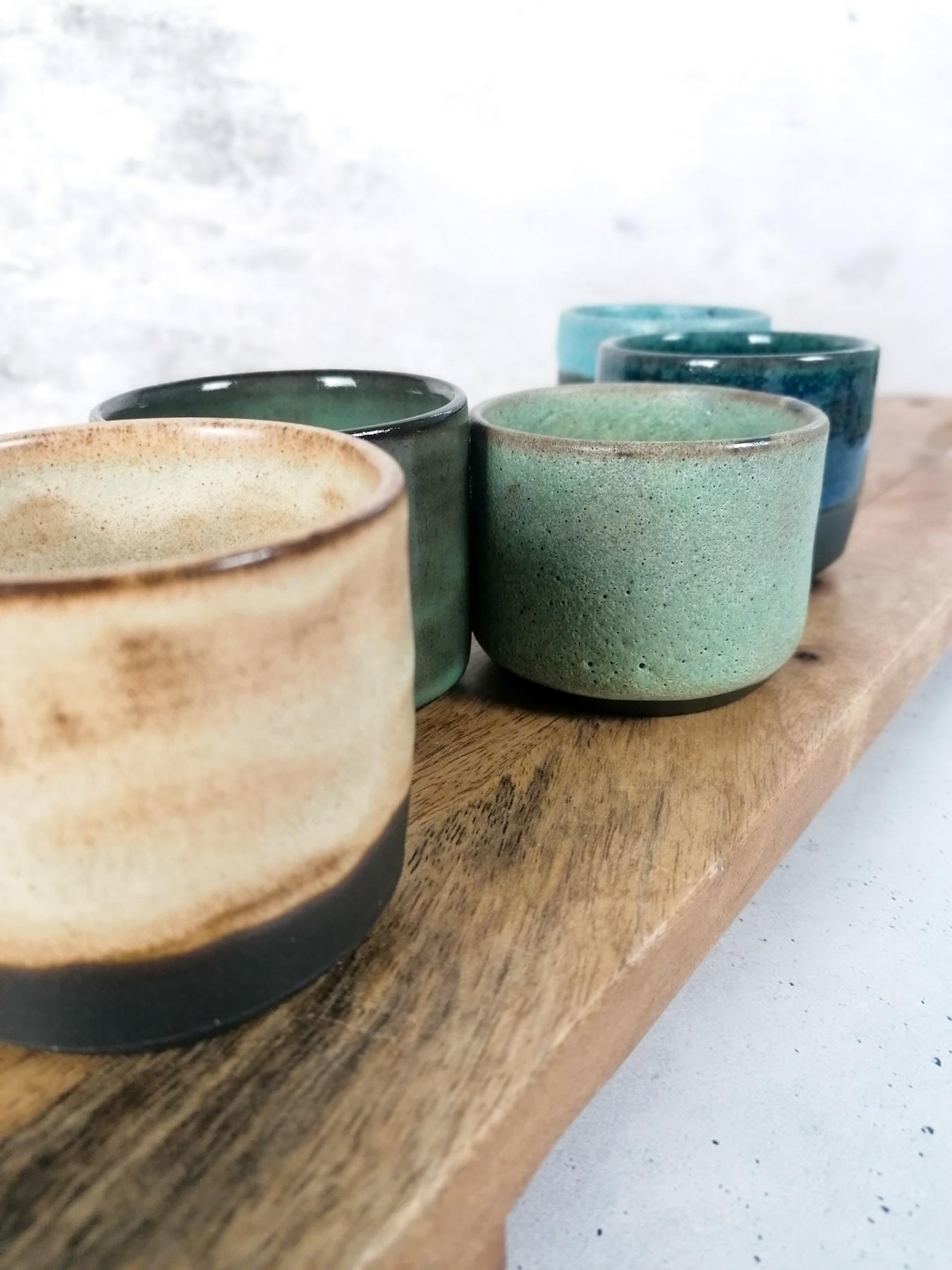 Sechs Botz Steinzeugbecher mit erdiger und türkisfarbener Glasur sind in zwei Reihen auf einem Holzbrett vor einem hellen, strukturierten Hintergrund angeordnet. Brennbereich: 1220-1280°C.