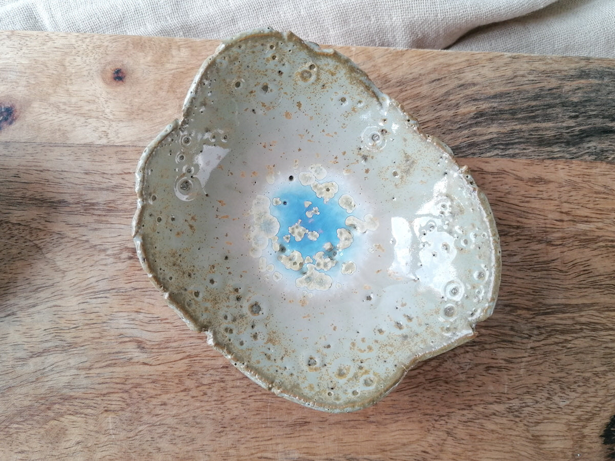 Die Botz Plus ist eine handgefertigte Keramikschale mit unregelmäßiger Form und glänzender, gesprenkelter Glasur. Die Steinzeug-Glasuren bilden ein einzigartiges blaues Kristallzentrum, während cremefarbene und braune Sprenkel die äußeren Bereiche zieren und für ein elegantes Aussehen sorgen.