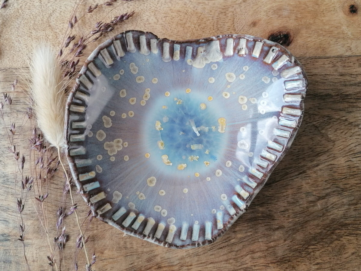 Eine handgefertigte, herzförmige Keramikschale mit glänzender blauer und beiger Botz Plus-Glasur steht auf einer Holzoberfläche, links davon sind getrocknete Gräser.