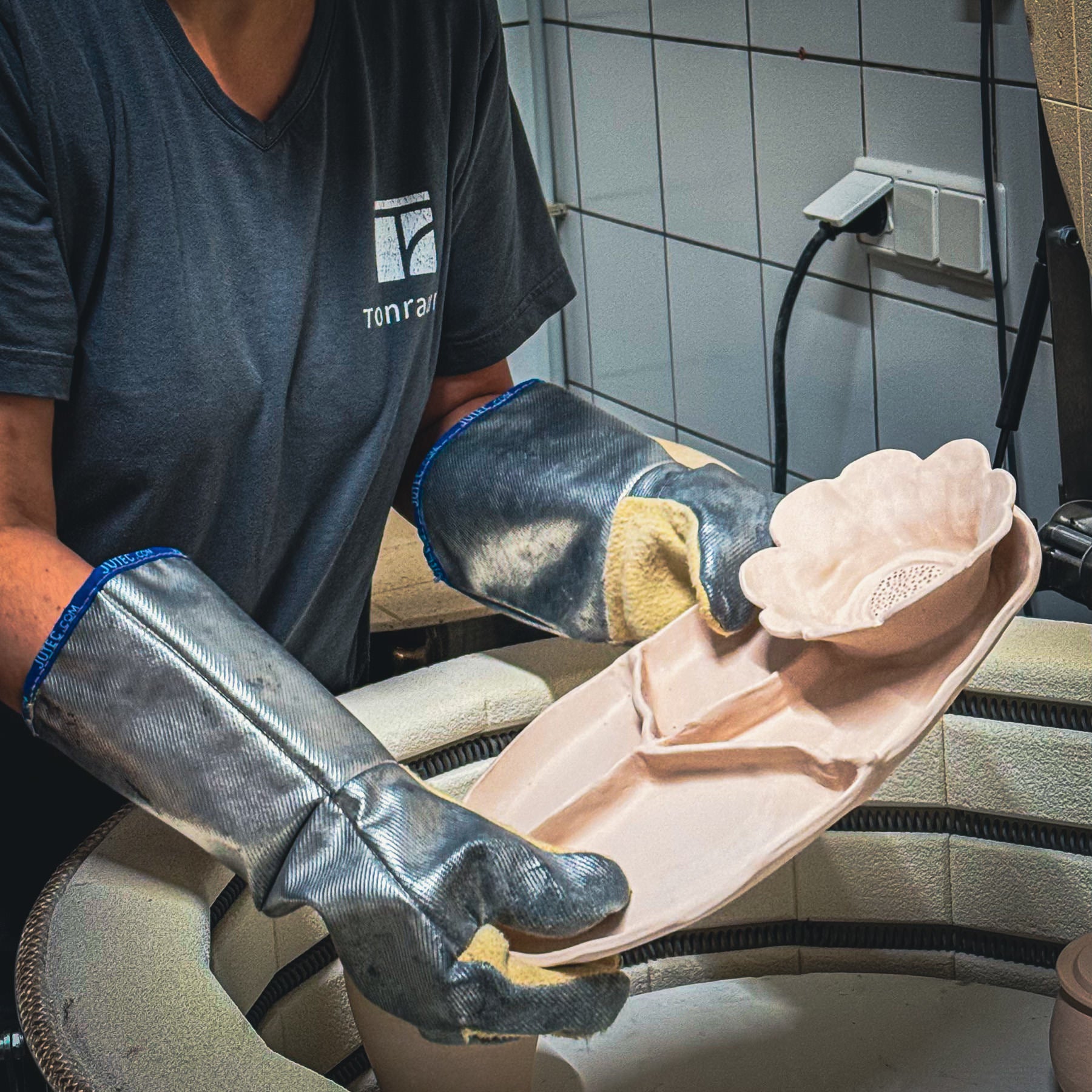 Eine Person mit silbernen hitzebeständigen Handschuhen hält eine Keramikplatte mit einem blumenförmigen Ausschnitt und steht neben einem Brennofen in einem gekachelten Raum.