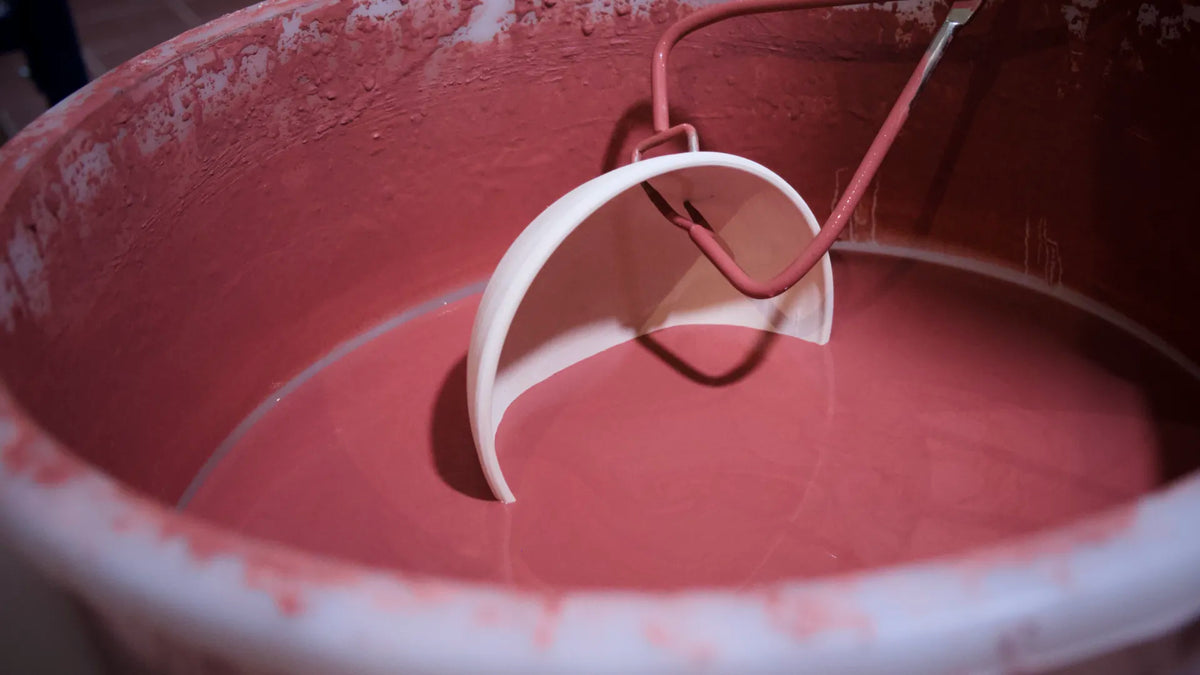 Im Kurs „Grundlagen des Glasierens“ wird eine weiße Keramikschale teilweise in einen großen Behälter mit rosa Glasur getaucht, wobei ihre Oberfläche mit Glasur überzogen wird, während ein Metallwerkzeug das Stück bei dieser klassischen Demonstration des Glasurauftrags festhält.