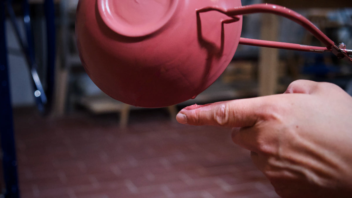 Eine Hand berührt behutsam eine rosa, nach oben gewendete Keramikschale, die an einem Metallhalter hängt, im Hintergrund ist ein verschwommener Ziegelboden zu sehen – ein Bild, das den zarten Moment des Glasierens einfängt, wie er im Kurs „Grundlagen des Glasierens“ vermittelt wird.
