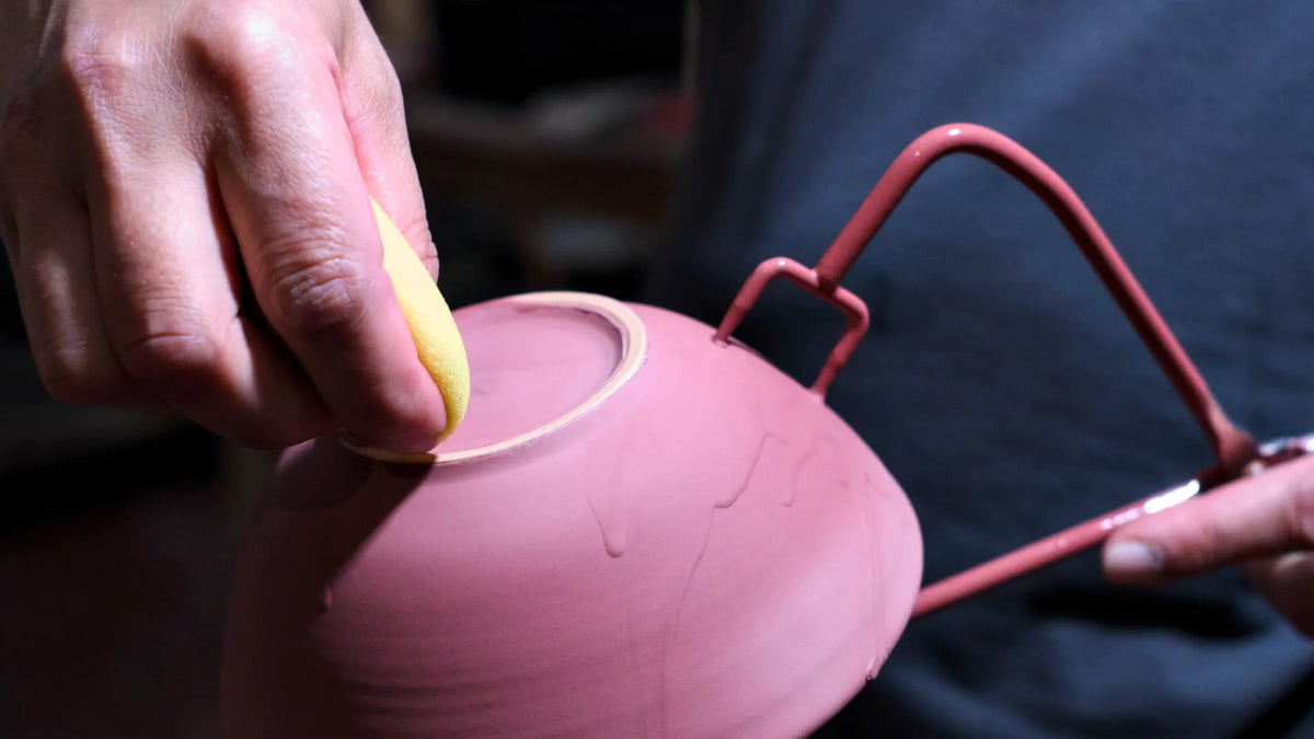 Jemand hält den Boden einer rosafarbenen, frisch glasierten Keramikschale mit einem Metallwerkzeug fest und glättet die Oberfläche mit einem gelben Schwamm – damit demonstriert er die sorgfältigen Techniken aus dem Kapitel „Grundlagen des Glasierens“, die häufig in einem Töpferkurs geübt werden.