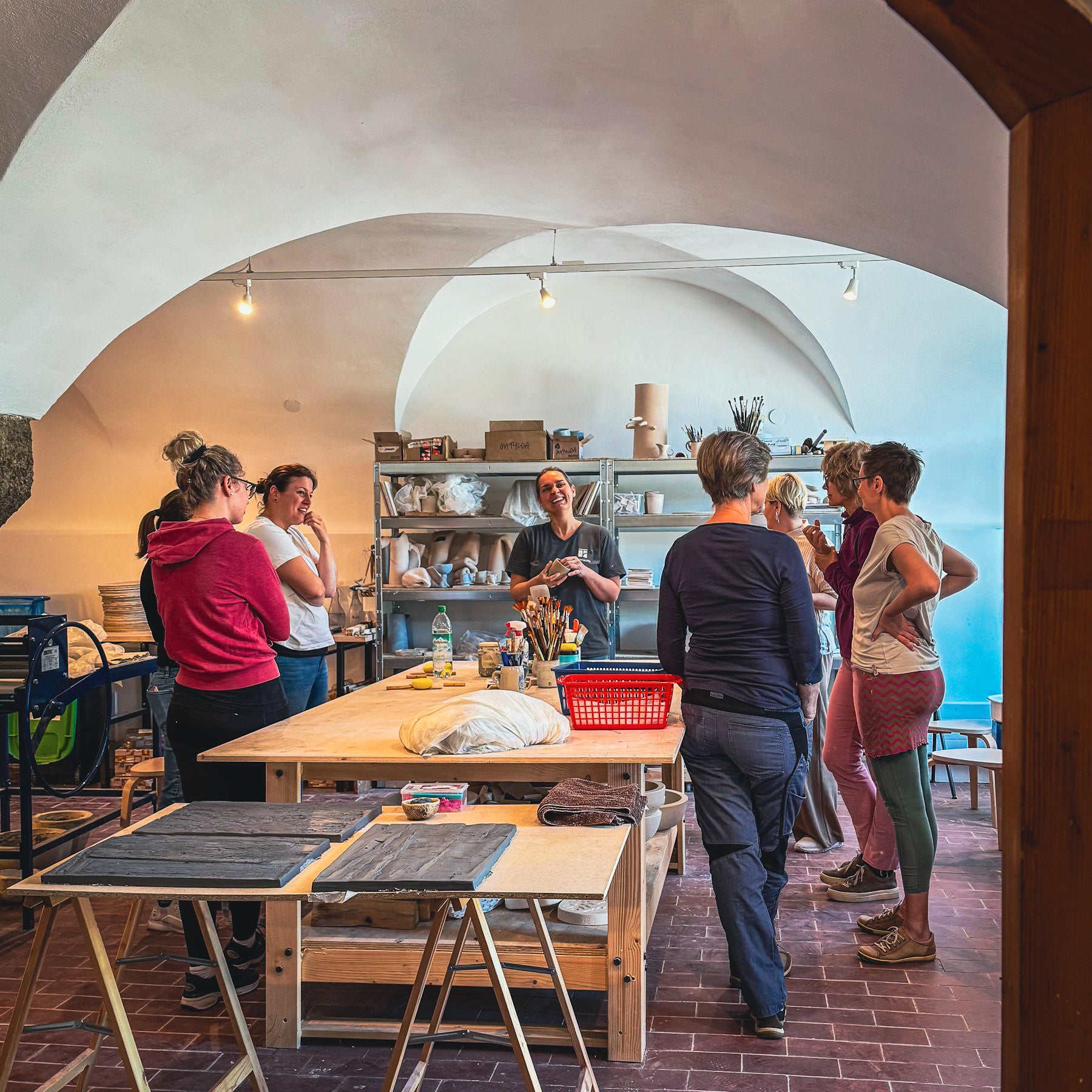 In einem hellen Töpferatelier steht eine Gruppe von Menschen um einen großen Tisch herum und hört einem Ausbilder zu. An der Rückwand stehen Regale mit Keramiken und Kunstzubehör. Der Raum hat gewölbte Decken und natürliches Licht.