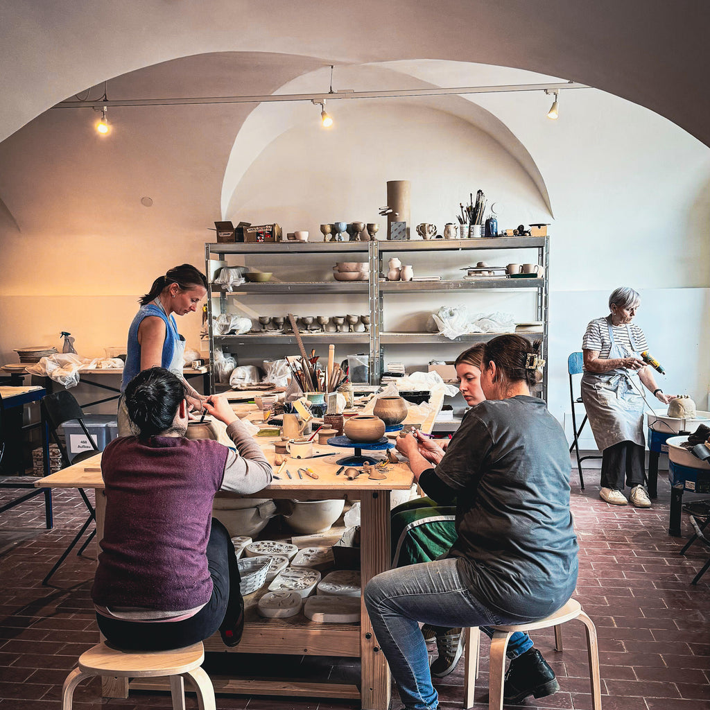 Vier Personen arbeiten an einem großen Holztisch in einem Keramikstudio, das mit Regalen voller Tonstücke und Werkzeuge gefüllt ist. Das Monatsabo umfasst Studio-Nutzung und Brennservice und schafft einen warmen, kreativen Raum für gemeinsame Projekte.