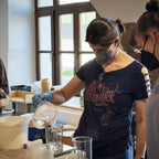 Eine Frau mit Mundschutz und Handschuhen gießt eine Flüssigkeit aus einem Messbecher in einen Becher, umgeben von Laborgeräten - sie demonstriert die präzise Glasurherstellung im Kurs "Glasuren Intensiv".