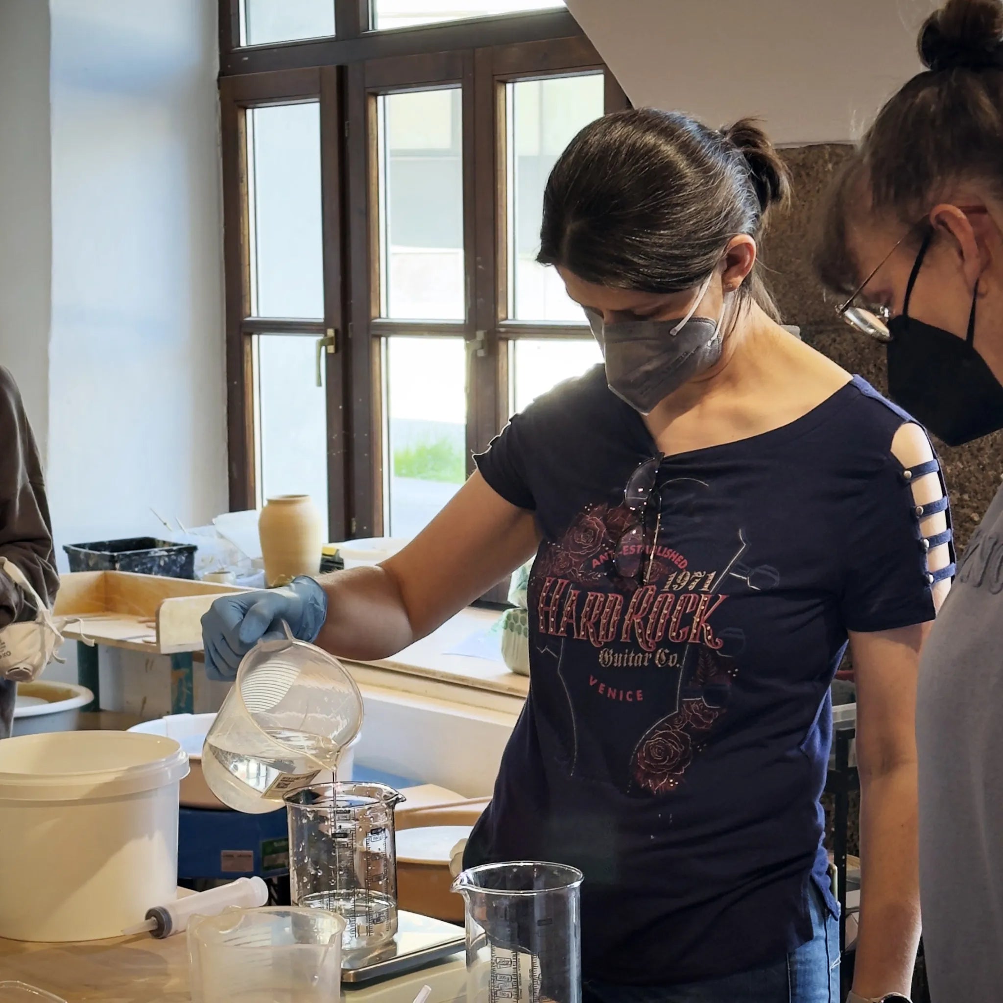 Eine Frau mit Mundschutz und Handschuhen gießt eine Flüssigkeit aus einem Messbecher in einen Becher, umgeben von Laborgeräten - sie demonstriert die präzise Glasurherstellung im Kurs "Glasuren Intensiv".