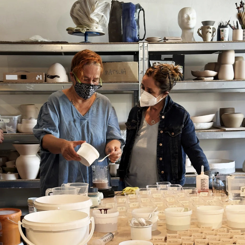 Zwei Frauen mit Gesichtsmasken arbeiten zusammen in einer Keramikwerkstatt, umgeben von Töpferwaren. Die eine gießt während der Zubereitung von Glasuren Instensiv Flüssigkeit ein, während die andere zuschaut. Im Hintergrund sind Regale zu sehen, die mit Keramikstücken gefüllt sind.