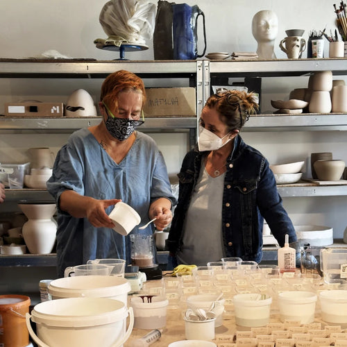 Zwei Frauen mit Gesichtsmasken arbeiten zusammen in einer Keramikwerkstatt, umgeben von Töpferwaren. Die eine gießt während der Zubereitung von Glasuren Instensiv Flüssigkeit ein, während die andere zuschaut. Im Hintergrund sind Regale zu sehen, die mit Keramikstücken gefüllt sind.