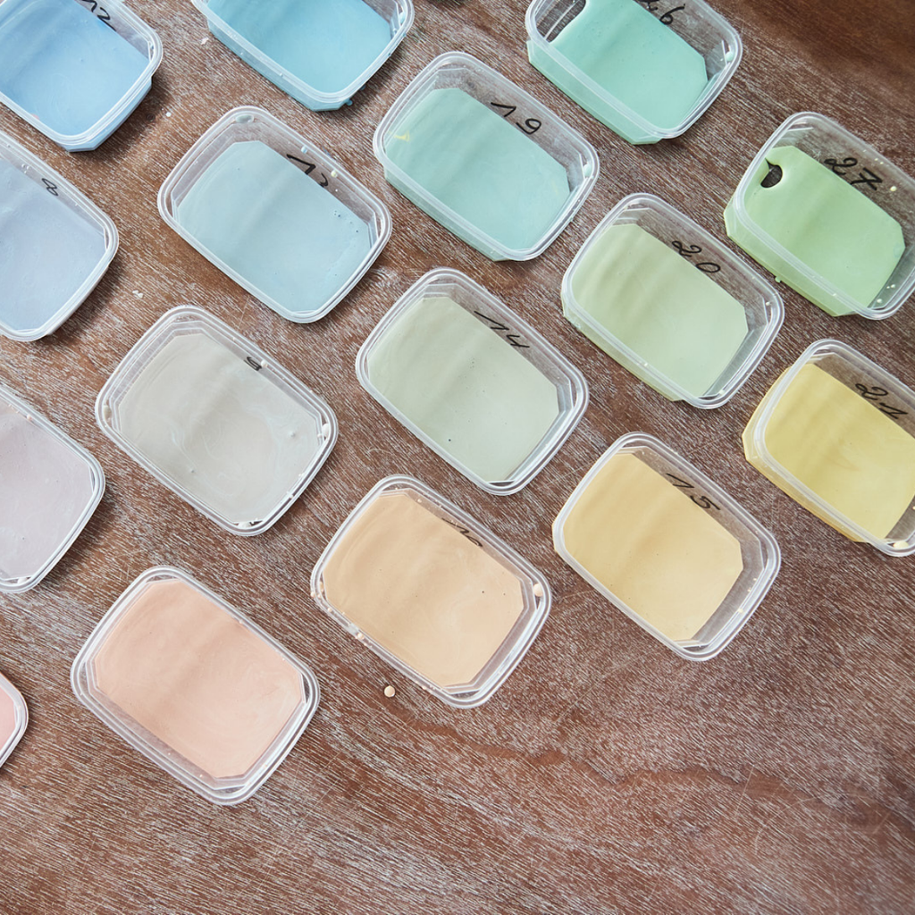 Kleine Plastikbehälter mit Pastellfarben (blau, grün, gelb, pfirsich) aus "Glasuren verstehen mit Sofie Goethals" sind ordentlich auf einer Holzfläche angeordnet - ideal für Glasurrezeptexperimente oder Ihren nächsten Glasur-Workshop. Auf einigen Deckeln sind handschriftliche Nummern angebracht.