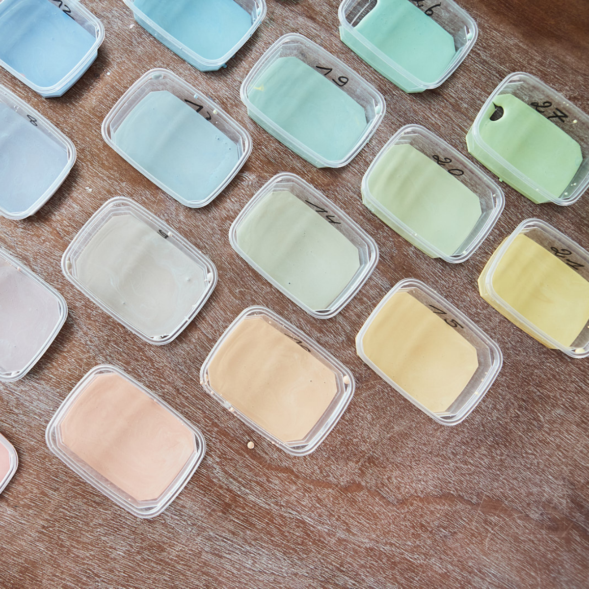 Kleine Plastikbehälter mit Pastellfarben (blau, grün, gelb, pfirsich) aus "Glasuren verstehen mit Sofie Goethals" sind ordentlich auf einer Holzfläche angeordnet - ideal für Glasurrezeptexperimente oder Ihren nächsten Glasur-Workshop. Auf einigen Deckeln sind handschriftliche Nummern angebracht.