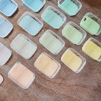 Kleine Plastikbehälter mit Pastellfarben (blau, grün, gelb, pfirsich) aus "Glasuren verstehen mit Sofie Goethals" sind ordentlich auf einer Holzfläche angeordnet - ideal für Glasurrezeptexperimente oder Ihren nächsten Glasur-Workshop. Auf einigen Deckeln sind handschriftliche Nummern angebracht.