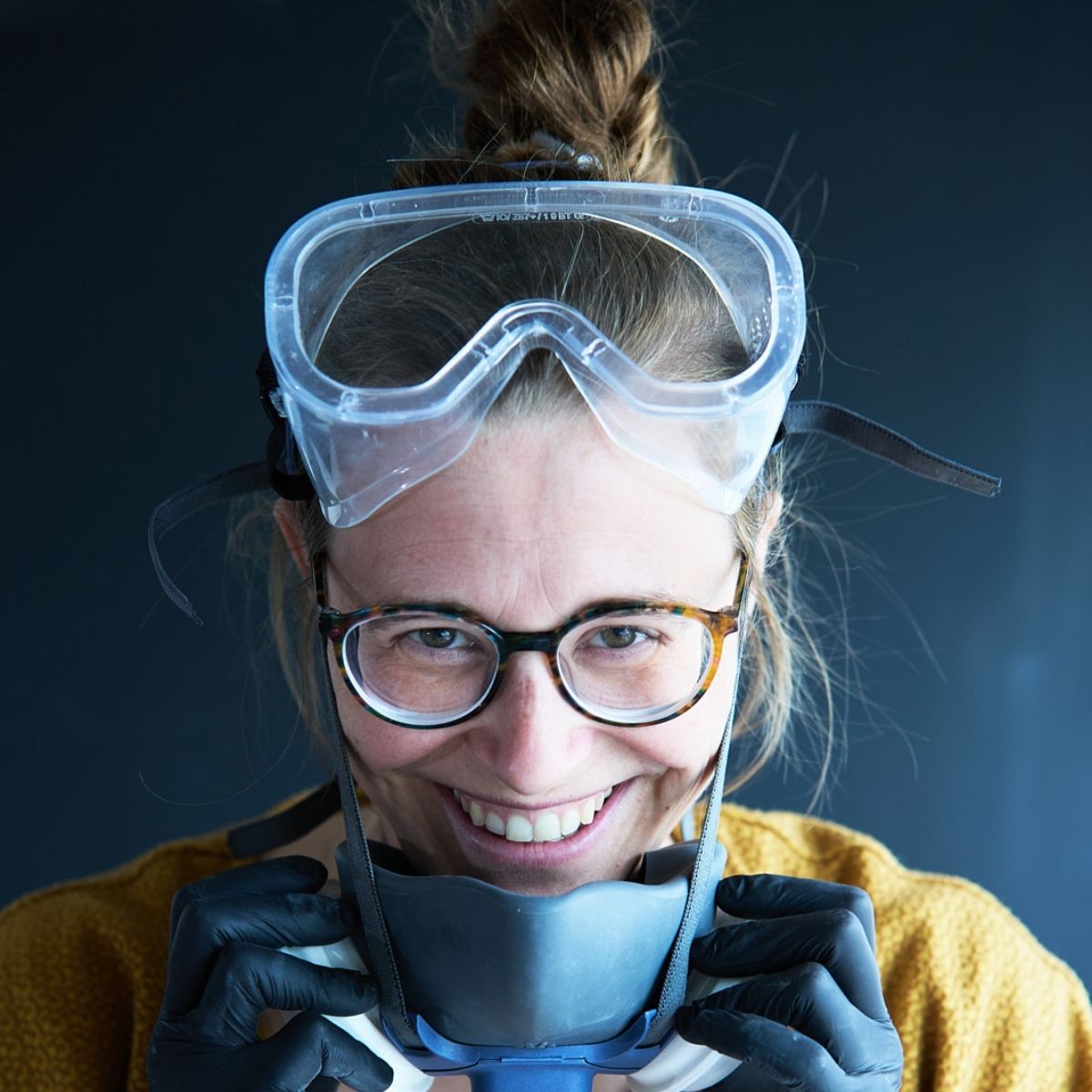 Mit schwarzen Handschuhen, Brille und einer Gesichtsmaske unter dem Kinn steht eine lächelnde Person bereit, bei „Glasuren verstehen mit Sofie Goethals“ vor dunklem Hintergrund mit neuen Glasurrezepten zu experimentieren.