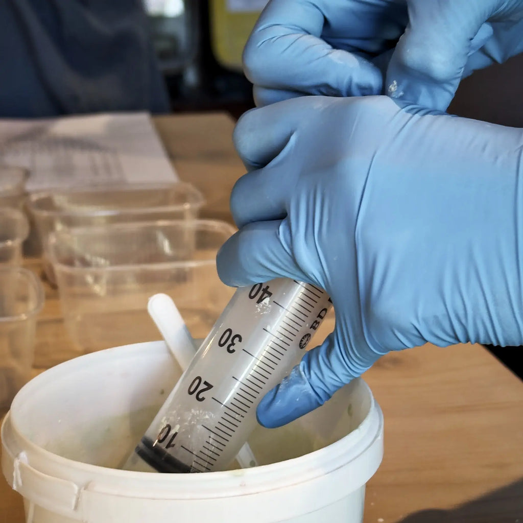 Eine Person mit blauen Handschuhen entnimmt mit einer großen Spritze Flüssigkeit für Glasuren Intensiv aus einem weißen Behälter auf einem Holztisch, in dessen Nähe sich kleine Plastikbehälter und Papier befinden - möglicherweise während eines Glasurkurses.