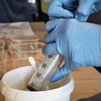 Eine Person mit blauen Handschuhen entnimmt mit einer großen Spritze Flüssigkeit für Glasuren Intensiv aus einem weißen Behälter auf einem Holztisch, in dessen Nähe sich kleine Plastikbehälter und Papier befinden - möglicherweise während eines Glasurkurses.