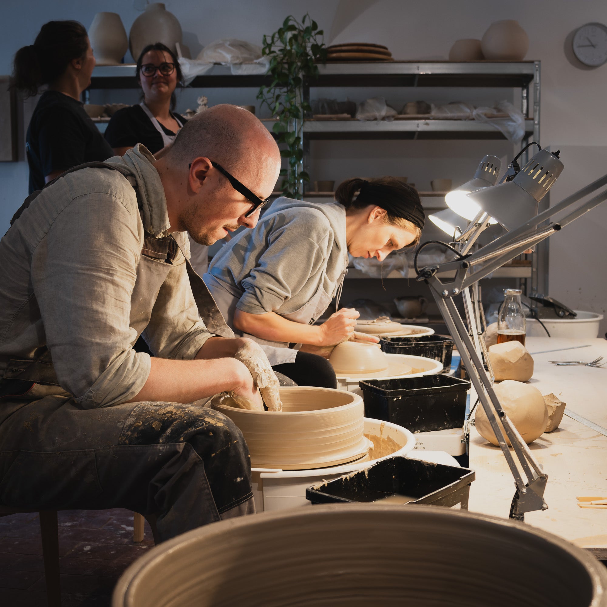 Zwei Personen formen in einem hellen Atelier Ton auf Töpferscheiben, benutzen Werkzeuge und Töpferwaren. Zwei andere schauen zu und lächeln. Regale mit Keramiken, eine Pflanze und Monatsabo-Details vervollständigen die Szenerie.