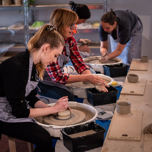 Vier Frauen mit Schürzen formen in einem Atelier Ton auf Töpferscheiben. Sie wirken konzentriert und in den kreativen Prozess vertieft, umgeben von Werkzeugen und fertigen Stücken auf Holztischen.