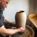 Ein bärtiger Mann mit einer Tätowierung übt das Zentrieren von Ton, indem er in einem hellen Atelier während des Meisterkurses eine hohe Vase auf einer Töpferscheibe formt: Höher drehen mit Oliver.
