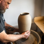 Ein bärtiger Mann mit einer Tätowierung übt das Zentrieren von Ton, indem er in einem hellen Atelier während des Meisterkurses eine hohe Vase auf einer Töpferscheibe formt: Höher drehen mit Oliver.