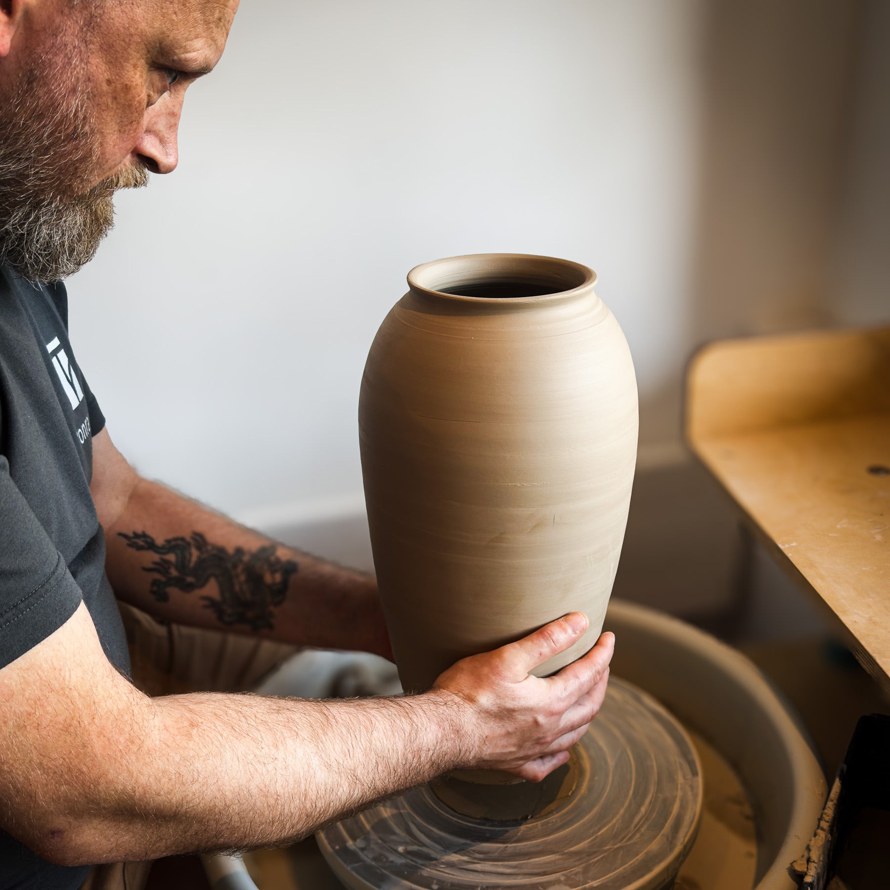 Ein bärtiger Mann mit einer Tätowierung übt das Zentrieren von Ton, indem er in einem hellen Atelier während des Meisterkurses eine hohe Vase auf einer Töpferscheibe formt: Höher drehen mit Oliver.