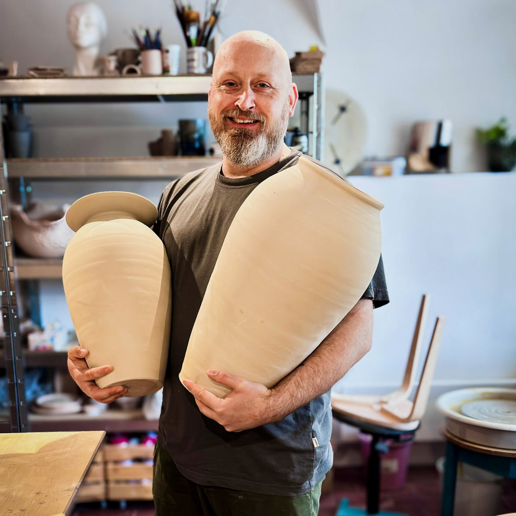 Ein bärtiger Mann lächelt in einer Töpferwerkstatt und hält zwei große, unfertige Vasen in der Hand. Hinter ihm stehen Regale mit Töpferwerkzeugen und spiegeln die kreative Atmosphäre von "Masterclass: Höher drehen mit Oliver" bei Zentrieren von Ton.
