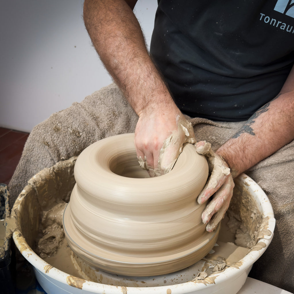 Während der "Masterclass: Höher drehen mit Oliver" formt ein Teilnehmer nassen Ton auf einem Spinnrad und zentriert ihn mit beiden Händen. Ihre Arme sind mit Ton bedeckt und ein Handtuch wird zum Schutz über ihren Schoß gelegt.