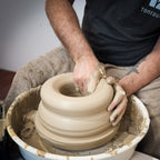 Während der "Masterclass: Höher drehen mit Oliver" formt ein Teilnehmer nassen Ton auf einem Spinnrad und zentriert ihn mit beiden Händen. Ihre Arme sind mit Ton bedeckt und ein Handtuch wird zum Schutz über ihren Schoß gelegt.