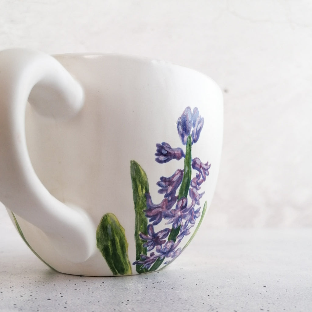 Eine weiße Keramiktasse mit geschwungenem Henkel, bemalt mit Lavendel und grünen Blättern mit Botz Unidekor, steht auf einer hellen Fläche vor einem schlichten Hintergrund – ideal für kreative Keramikprojekte.