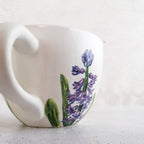 Eine weiße Keramiktasse mit geschwungenem Henkel, bemalt mit Lavendel und grünen Blättern mit Botz Unidekor, steht auf einer hellen Fläche vor einem schlichten Hintergrund – ideal für kreative Keramikprojekte.