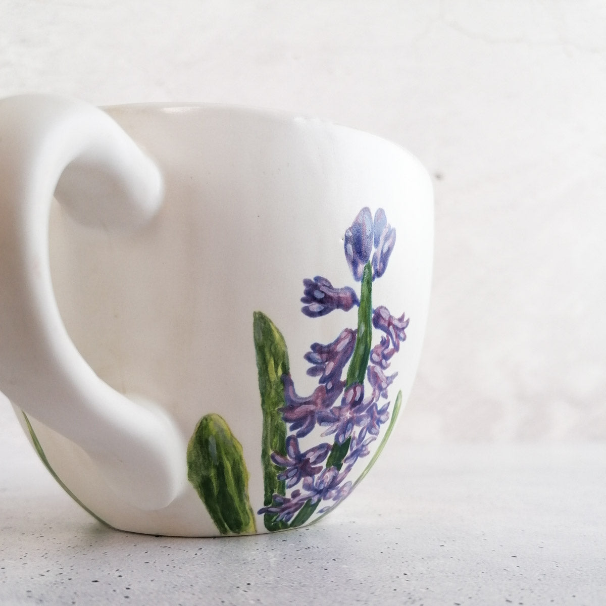 Eine weiße Keramiktasse mit geschwungenem Henkel, bemalt mit Lavendel und grünen Blättern mit Botz Unidekor, steht auf einer hellen Fläche vor einem schlichten Hintergrund – ideal für kreative Keramikprojekte.