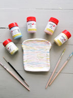 Fünf kleine Gläser mit Pastellfarben von Botz Unidekor umgeben einen gewellten, toastförmigen Keramikteller mit drei farbigen Pinseln – die perfekte Inspiration für Ihr nächstes Keramikprojekt.