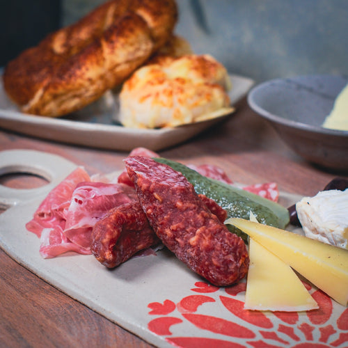 Auf dem "Gestalte dein eigenes Jausenbrett & Dipschale aus Ton" wird ein Wurstbrett mit Wurst, Salami, Käse und einer Essiggurke serviert, daneben liegen auf einem Holztisch Kekse und extra Käse. Das Set verleiht dem Arrangement eine schöne Note.