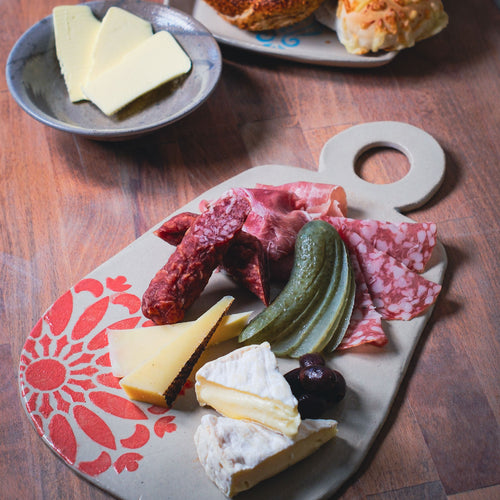 Auf dem Holztisch "Gestalte dein eigenes Jausenbrett & Dipschale aus Ton" ist neben Butter und Brötchen ein Wurstbrett mit Wurst, Käse, einer Gurke und Oliven ausgestellt.
