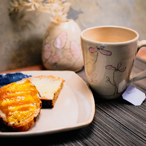 Das "Dein selbstgemachtes Frühstücks-Set aus Ton" umfasst eine Keramiktasse mit Blumenmuster, einen Teller mit zwei Brotscheiben (Butter und Orangenmarmelade) und eine kleine Vase mit Trockenblumen auf einem gestreiften Platzset.