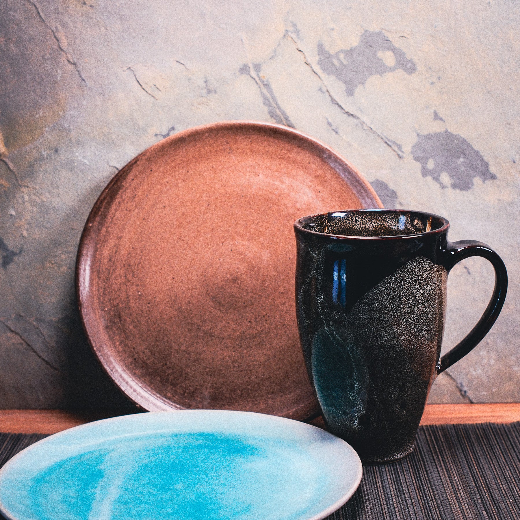 Ein brauner Keramikteller, ein hoher dunkler Keramikbecher und ein weißer Teller mit blauem Zentrum liegen auf einer dunklen Unterlage-perfekt als Inspiration für Dein selbstgemachtes Frühstücks-Set aus Ton.