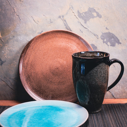 Ein brauner Keramikteller, ein hoher dunkler Keramikbecher und ein weißer Teller mit blauem Zentrum liegen auf einer dunklen Unterlage-perfekt als Inspiration für Dein selbstgemachtes Frühstücks-Set aus Ton.