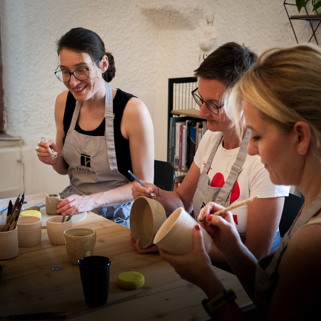 In einem Tonraum-Kaffeekränzchen-Workshop bemalen drei Frauen lächelnd Keramikbecher und -schalen an einem Holztisch, umgeben von Malutensilien und Pinseln, in einer kreativen, entspannten Atmosphäre.