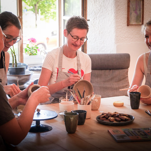 Im Tonraum Kaffeekränzchen lächeln drei Frauen, während sie gemeinsam Töpferwaren bemalen, umgeben von Kunstzubehör, Snacks und Kaffeespezialitäten oder Tee auf einem Holztisch. Durch das Fenster dringt Sonnenlicht herein und sorgt für eine gemütliche, fröhliche Atmosphäre.